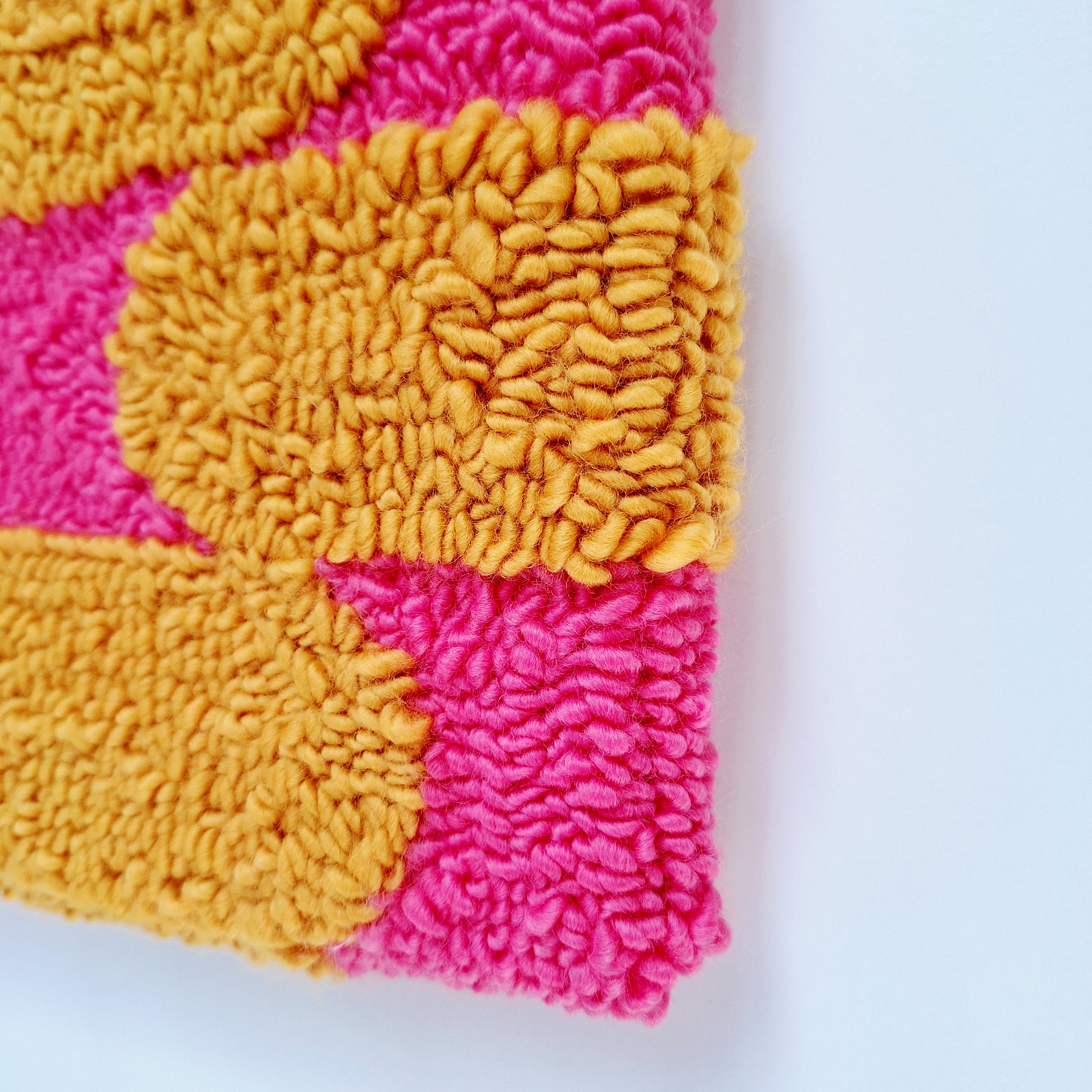Vista de cerca de cuadro de lana realizado artesanalmente en punch needle. Se aprecia relieve de bordado. Color rosa y mostaza.