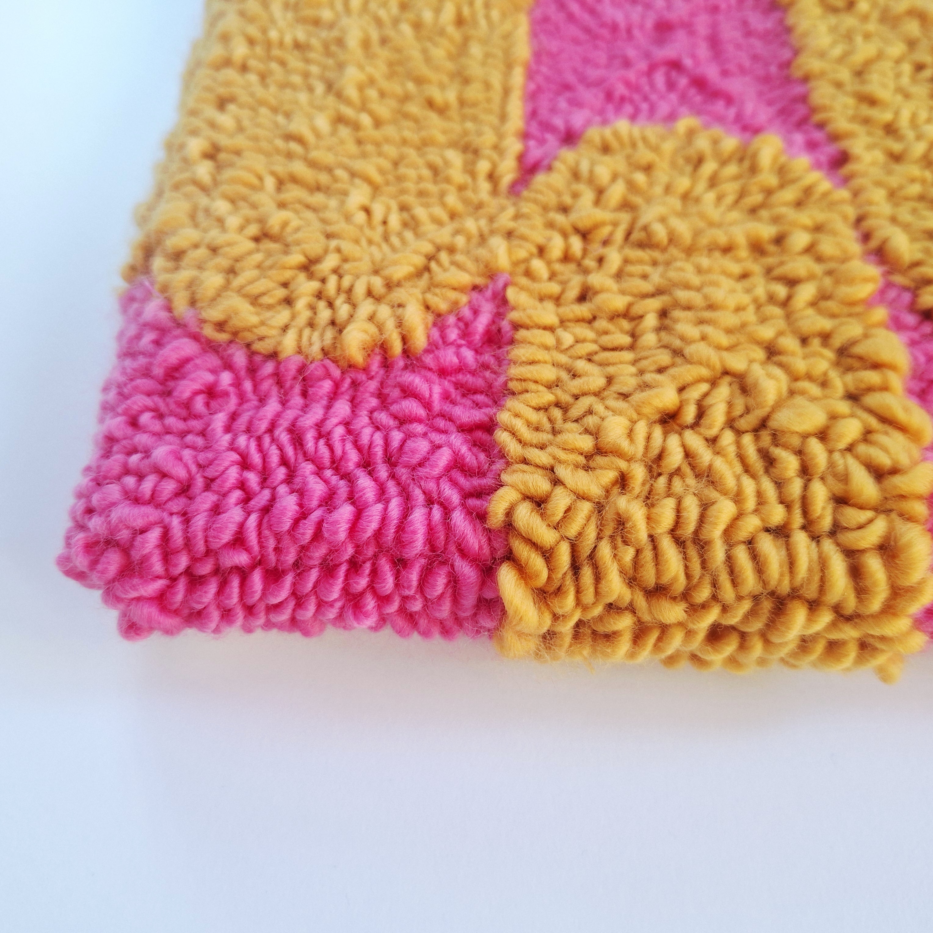 Vista de cerca de cuadro de lana realizado artesanalmente en punch needle. Se aprecia relieve de bordado. Color rosa y mostaza.